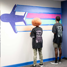 Load image into Gallery viewer, Two people paint green and orange lines under blue and pink lines trailing behind an airplane at the start of the macon airport mural
