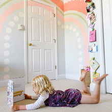 Load image into Gallery viewer, Final rainbow mural in B's bedroom. The rainbow is different shades of pink and a stripe of sage green. The outermost stripe is salmon pink and has scalloped edges. The second stripe is about an 1in thick and is sage green. The third stripe is a mid tone pink about 3in wide.The last stripe is light pink circles. There are pink clouds near the rainbow and a little girl laying on the floor reading a book below the rainbow.