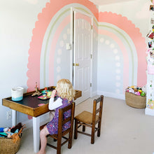 Load image into Gallery viewer, Final rainbow mural in B's bedroom. The rainbow is different shades of pink and a stripe of sage green. The outermost stripe is salmon pink and has scalloped edges. The second stripe is about an 1in thick and is sage green. The third stripe is a mid tone pink about 3in wide.The last stripe is light pink circles. There are pink clouds near the rainbow and a little girl is sitting at a handmade table having a tea party below the rainbow.
