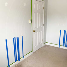 Load image into Gallery viewer, Initial set up for B's rainbow mural. The rainbow lines are chalked out and painter's tape is on the wall and around the baseboards and door frame.