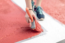 Load image into Gallery viewer, Woman's hand holding a paint brush with red paint on the corned of taped crosswalk