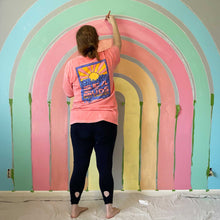 Load image into Gallery viewer, Abby works on the second coat for E's rainbow mural. She's painting the warm pink stripe and you can also see the first coats of the sky blue, soft green, salmon pink, creamy orange, and buttery yellow stripes. Between each stripe is a smaller stripe of the dark gray wall.