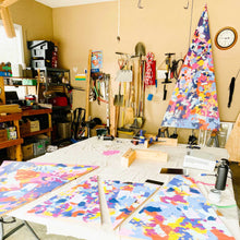 Load image into Gallery viewer, In the middle of painting and creating the four holiday trees for the Museum of Arts and Sciences Macon. On the work table you'll see four smaller trees covered in splashes of warm and bright colors. In the background is the main tree, also covered in abstract lines and dots in bright colors.