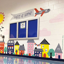 Load image into Gallery viewer, Ground view of "habits at home" mural with multicolored houses and buildings at the bottom of the wall and an airplane holding a banner with "habits at home" hand-lettered in black text.
