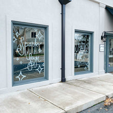 Load image into Gallery viewer, Close-up of two glass windows separated by a pole on a building. The windows have green diamond shapes and dots; the left window has "Sparks Yoga" painted in white, and the right window has "what sparks your soul?" painted in white paint.