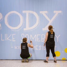 Load image into Gallery viewer, Close up of two women painting white letters on the Blue wall mural with white text that says "be someone who makes everybody feel like somebody"-kid president"