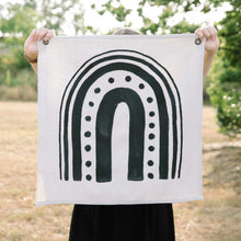 Load image into Gallery viewer, Erin from Mama Hawk Draws holds a 2'x2' cream colored drop cloth with a black rainbow painted on it. The rainbow has different widths of stripes. The interior stripe is the thickest, about 3", the next stripe is made of medium sized circles, the third stripe is about 2" thick, the 4th stripe is about 1" thick, and the last stripe is about 0.5" thick.