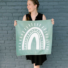 Load image into Gallery viewer, Erin from Mama Hawk Draws holds a 2'x2' green colored drop cloth with a white rainbow painted on it. The rainbow's four stripes have different thicknesses with one stripe made of dashes. Outside the stripes are rays around the rainbow.