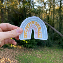 Load image into Gallery viewer, Hand holding a pink, yellow, and blue rainbow magnet with greenery in background