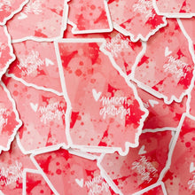 Load image into Gallery viewer, State of Georgia sticker with red and pink watercolor shaded pattern. Small white heart and text "macon, georgia" off right center of the sticker, and the background is a collage of the same sticker