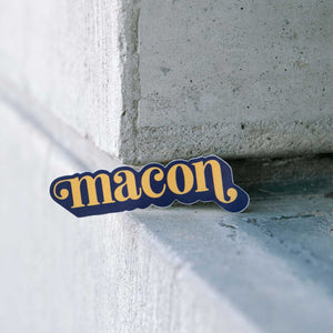 Sticker sitting on a concrete ledge says "Macon" in orange inlower case serif font with ball flourishes on the "m" and the descender of the "n." The word has a angled drop shadow in navy.