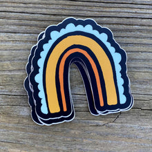 Load image into Gallery viewer, On a wooden table rests a stack of  3x3" rainbow stickers. The rainbow has three stripes. The inside one is a thin orange line, the middle one is a golden rod thicker line, and the outer stripe is a team scalloped line. The background color is black.