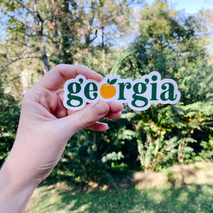 Woman's hand holding white sticker with "Georgia" text in green and orange peach as the "o" letter with greenery in the background