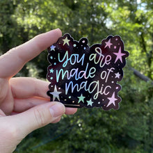 Load image into Gallery viewer, Woman holding a black sticker with colorful hologram hand lettering that reads "you are made of magic" surrounded by stars. The sticker is being held in front of greenery.