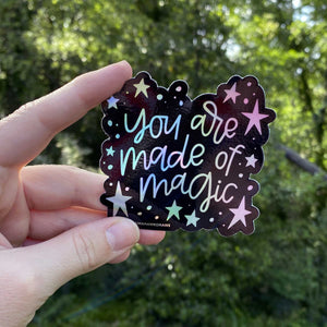 Woman holding a black sticker with colorful hologram hand lettering that reads "you are made of magic" surrounded by stars. The sticker is being held in front of greenery.