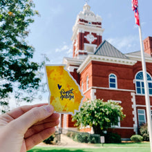 Load image into Gallery viewer, A white woman holds up a state of Georgia shaped sticker with a yellow abstract design in front of a building with a clocktower. Text in a curly font "forsyth, georgia" and a heart are layered on top of the sticker in dark blue.