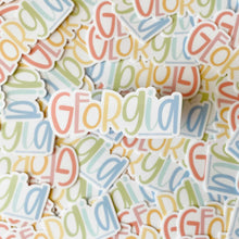 Load image into Gallery viewer, A sticker with text "Georgia". The G is red, E is a dim pink, O is orange, R is yellow, G is green, I is light blue, and A is blue. The background color is white. Behind the focused sticker is a pile of many of the same sticker.
