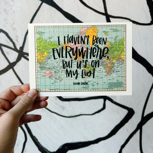 A horizontal multicolored vintage style map of the world with hand lettered text that says "I haven't been everywhere, but it's on my list" in black lettering. The map is held in front of a white backdrop with spiraling black lines by a white woman.