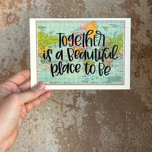 A blue, green, yellow, and pink flat map of the world with text "together is a beautiful place to be" in black lettering over the map print. The map is held in front of a brown rustic wall by a white woman.