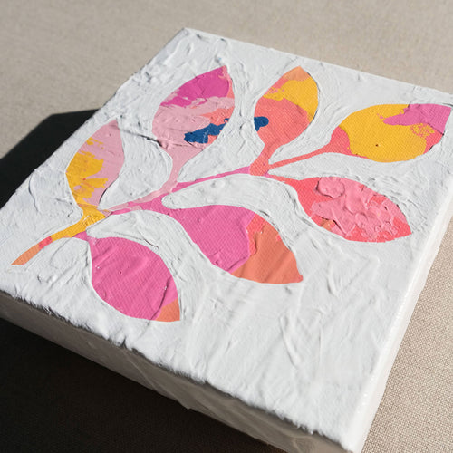 A colorful abstract painting with colors pink, yellow, orange, and some blue painted over with white to show a stem with leaves in the negative colored space. The painting is laying on a tan canvas.