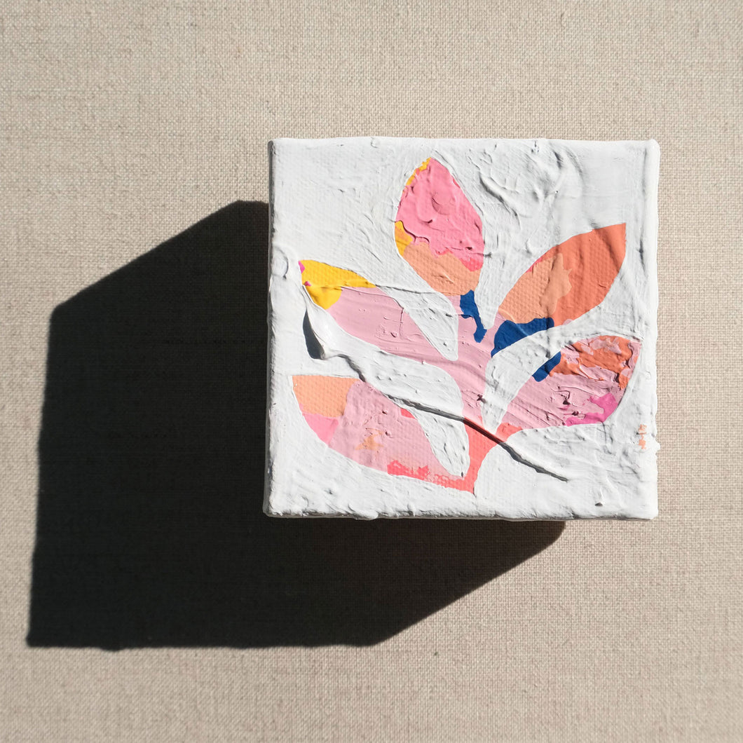 A colorful abstract painting with colors pink, yellow, orange, and some blue painted over with white to show a stem with leaves in the negative colored space. The painting is laying on a tan canvas.