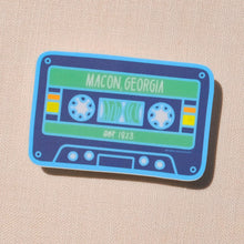 Load image into Gallery viewer, A stylized cassette tape design with colors light blue, navy blue, green, yellow, orange, and white. Text &quot;Macon, Georgia&quot; and &quot;est. 1823&quot; are printed in white on the design. The sticker is in front of a tan canvas.
