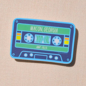 A stylized cassette tape design with colors light blue, navy blue, green, yellow, orange, and white. Text "Macon, Georgia" and "est. 1823" are printed in white on the design. The sticker is in front of a tan canvas.