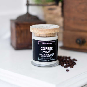 7th Street Salvage's Coffee Mill candle sitting on a white counter with a vintage coffee mill in the background. The candle label is black with white typography and the container has a wooden lid.