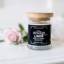 Load image into Gallery viewer, 7th Street Salvage&#39;s Vintage Linen candle sitting on a white counter with a pink rose n the background. The candle label is black with white typography and the container has a wooden lid.
