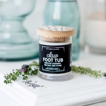 Load image into Gallery viewer, 7th Street Salvage&#39;s Claw Foot Tub candle sitting on a white counter with lavender sitting next to it. The candle label is black with white typography and the container has a wooden lid.
