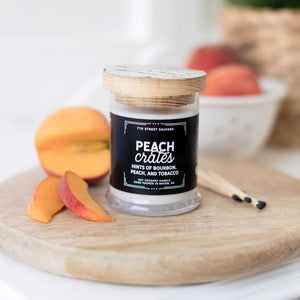 7th Street Salvage's Peach Crates candle sitting on a wooden cutting board with sliced peaches sitting next to it. The candle label is black with white typography and the container has a wooden lid.