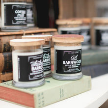 Load image into Gallery viewer, 7th Street Salvage&#39;s candles witting on vintage books. The books are green, blue, and brown. The candles labels are black with white typography and wooden lids. 
