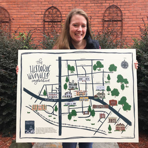 Mama Hawk Draws holding a painted map labeled "the historic vineville neighborhood" with black lines and painted buildings and trees, with brick and greenery in the background