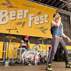 Photo of a band performing on a stage with living social beer fest sign enlarged in the stage background. The vocalist is standing center stage singing into a microphone, and a drummer is in the background playing drums