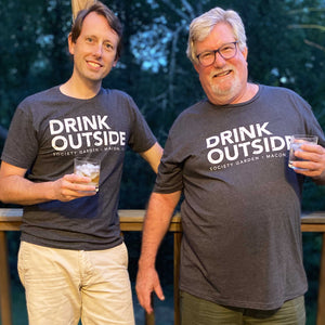 Two white men holding drinks with grey tees with "drink outside" printed in the center. They are leaning on a wooden rail with greenery in the background