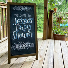 Load image into Gallery viewer, Stand up easel  chalkboard sign. At the top and bottom are handdrawn flowers in white. In the middle there is hand lettering that says &quot;welcome to&quot; is a monolone san serif and &quot;Jessie&#39;s Baby Shower&quot; in a hand lettered cursive all in white. 
