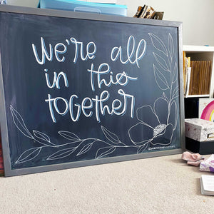 Larger Than Life's monthly chalkboard design. This month's board says "we're all in this together" in white lettering with a blue drop shadow. In the bottom right corner there is a flower with leaves.