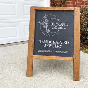 Hand drawn logo for Beyond the Stars on a small stand up easel. Below the crescent moon logo, it says "handcrafted jewelry" and social media information. The chalkboard background is black and all the lettering and illustrations are in white. 