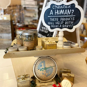 Chalkboard signage that is white framed with a black background. The lettering says, "Created a human? These products will help both Mom & baby!" in white. The framed chalkboard sits on top of a vintage baby scale amongst bath and beauty products. 