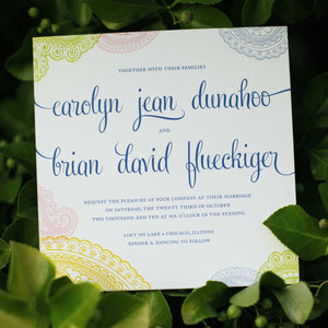 Photo of a multicolored card with cursive design text and mandala patterns on the edges. Wedding invitation with leaves in the background