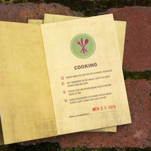 Load image into Gallery viewer, Yellow card open with green circle emblem of utensils. &quot;Cooking&quot; text in middle with checkboxes under and date with a stamp at the bottom. Brick background with moss in cracks
