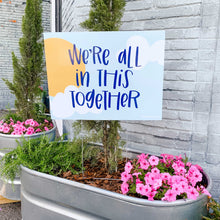 Load image into Gallery viewer, Photo of a yard sign sitting in a metal flower pot with greenery and pink flowers. The sign has a sky background with clouds and sun and &quot;we&#39;re all in this together&quot; handlettered in blue in the center
