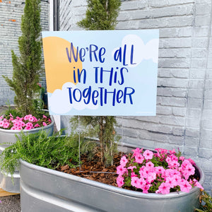 Photo of a yard sign sitting in a metal flower pot with greenery and pink flowers. The sign has a sky background with clouds and sun and "we're all in this together" handlettered in blue in the center