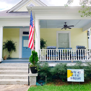 Street view of a yellow house with "we're all in this together" yard sign sitting in the grass in the bottom right.