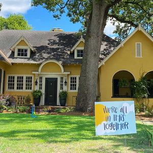 Street view of a yellow house with "we're all in this together" yard sign sitting in the grass in the bottom right. 
