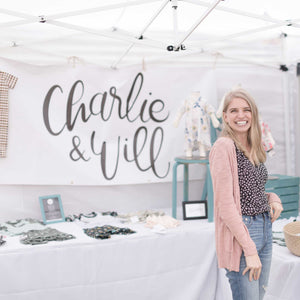 Owner, Kirsten McClendon, standing in front of her Charlie & Will booth were she is selling her handmade goods for littles. Behind Kirsten is a large white banner with the handlettered logo in black.