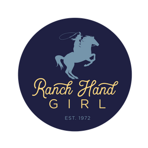 Navy blue circle logo with text "ranch hand girl" in yellow. "Ranch Hand" is in script lettering, and "girl" is in a rounded san serif font kerned out. Below the yellow type is "EST. 1972" in a smaller, light blue san serif font. Above all the type is a lighter blue horse on the back two hind legs with a female holding a lasso.