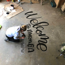 Load image into Gallery viewer, Mama Hawk Draws kneeling on concrete floor painting &quot;welcome to macon, GA&quot; with coordinates in black lettering
