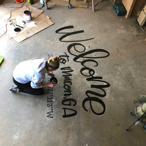Mama Hawk Draws kneeling on concrete floor painting "welcome to macon, GA" with coordinates in black lettering