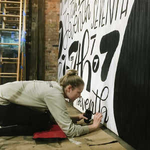 Mama Hawk Draws painting black lettering on the white wall of the 7th street salvage mural